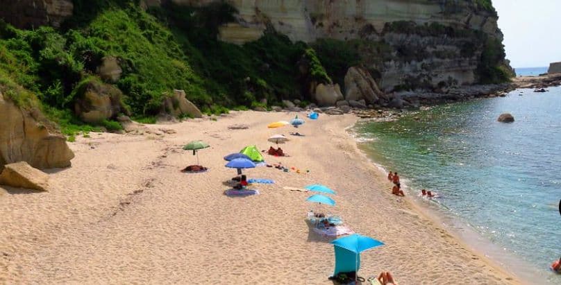 secluded white sandy beach with rocky cliffs and lush green vegetation behind, on a sunny day with a calm blue sea. several people under blue, yellow and green sun umbrellas, and 5 people at the waters edge bathing.