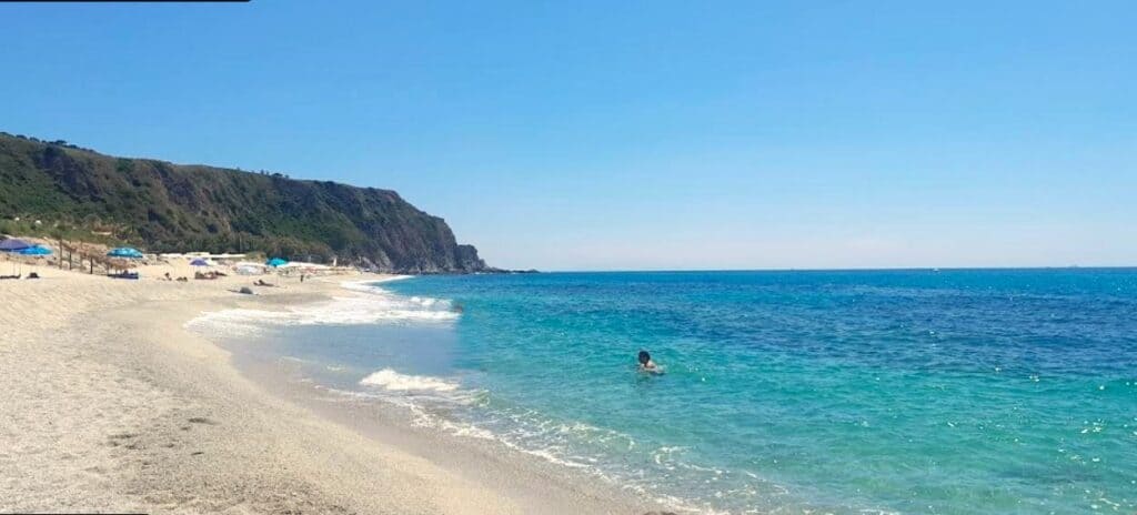 white sand beach, clear blue sea with person wading and green cliffs in the background, on a sunny day
