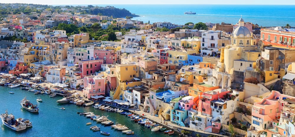 Aerial view of Procida Harbour, one of the Hidden Gems in Southern Italy. Pastel coloured houses with the boats moored and the blue sea in the background.
