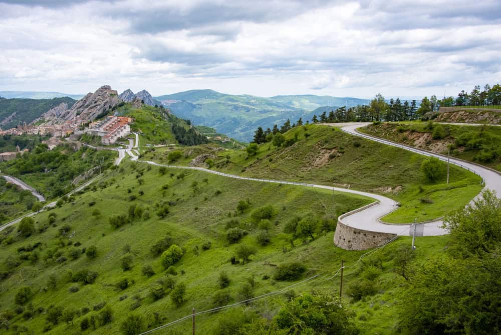 Historic village of Pietrapertosa in the distance, with lush green rolling hills and a narrow winding road in the foreground.