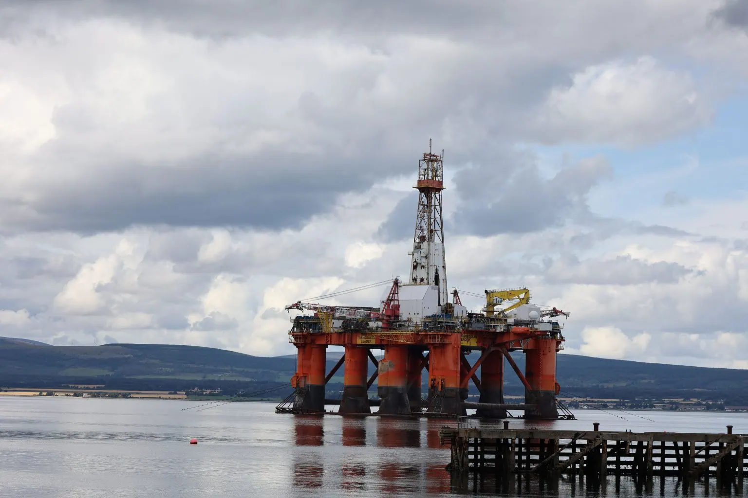 A large body of water dominated by an oil rig in the foreground and hillside in the back ground. An on old wooden jetty sits on the right in front of the oil rig.