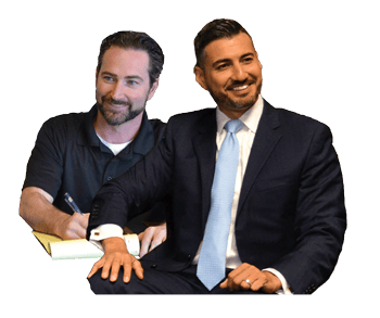 Two men are seated; one in a black shirt writing in a notebook, the other in a suit and tie, smiling.