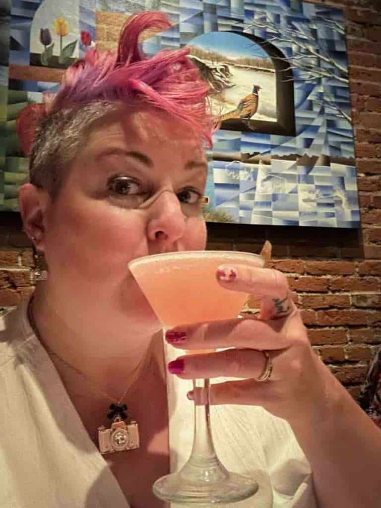 Pink-haired woman enjoying a cocktail at a bar with colorful artwork in the background.