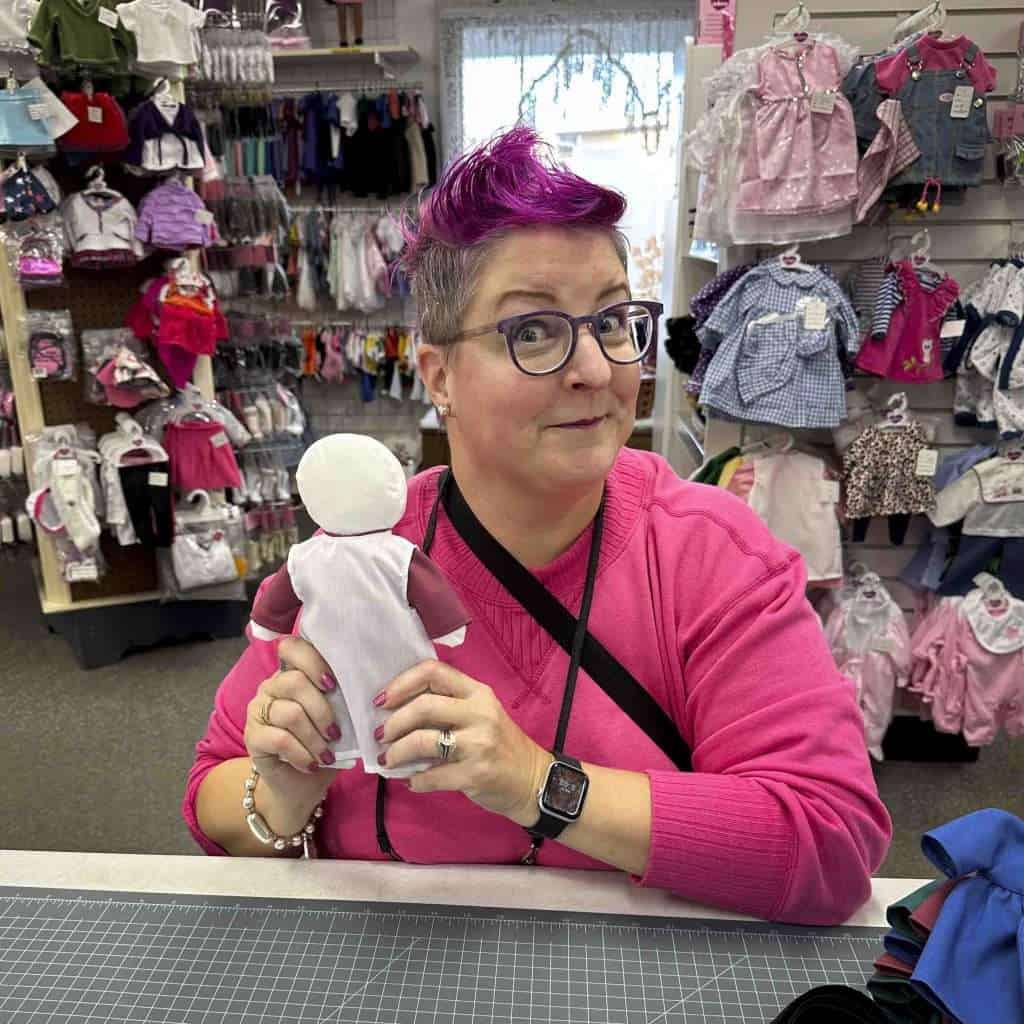 Pink-haired grandma holding a handmade doll in a children's clothing store, surrounded by small outfits and accessories, showcasing her vibrant personality and love for craft projects.