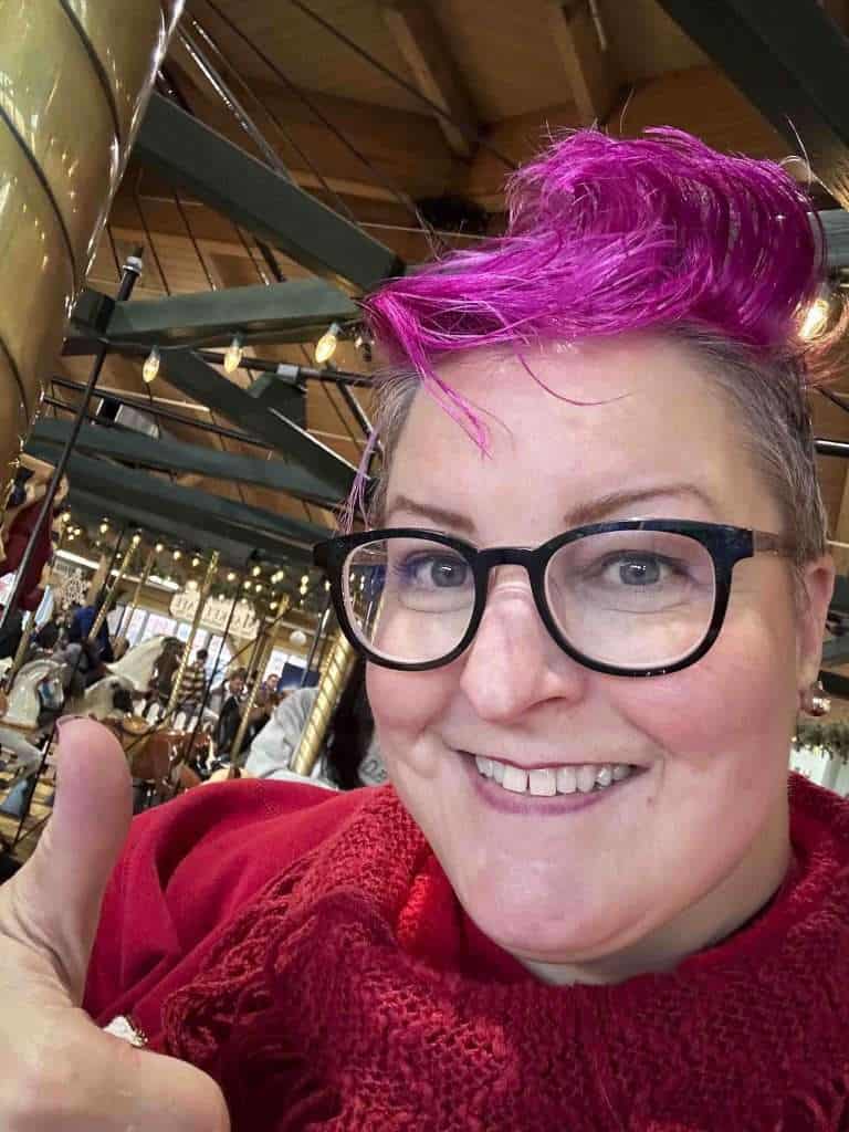 The pink-haired Hip Grandma gives a thumbs up while riding a carousel
