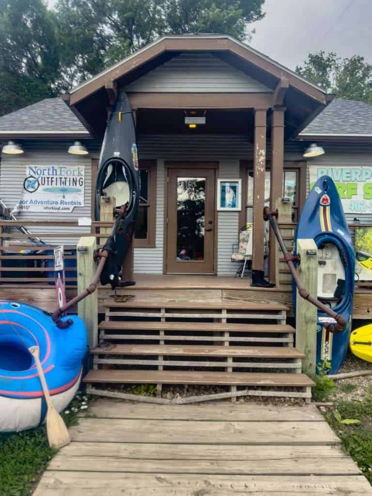 Entrance to North Fork Outfitting in Norfolk, Nebraska, showing kayaks, a blue rental tube, and a wooden porch with signage for outdoor adventure rentals.