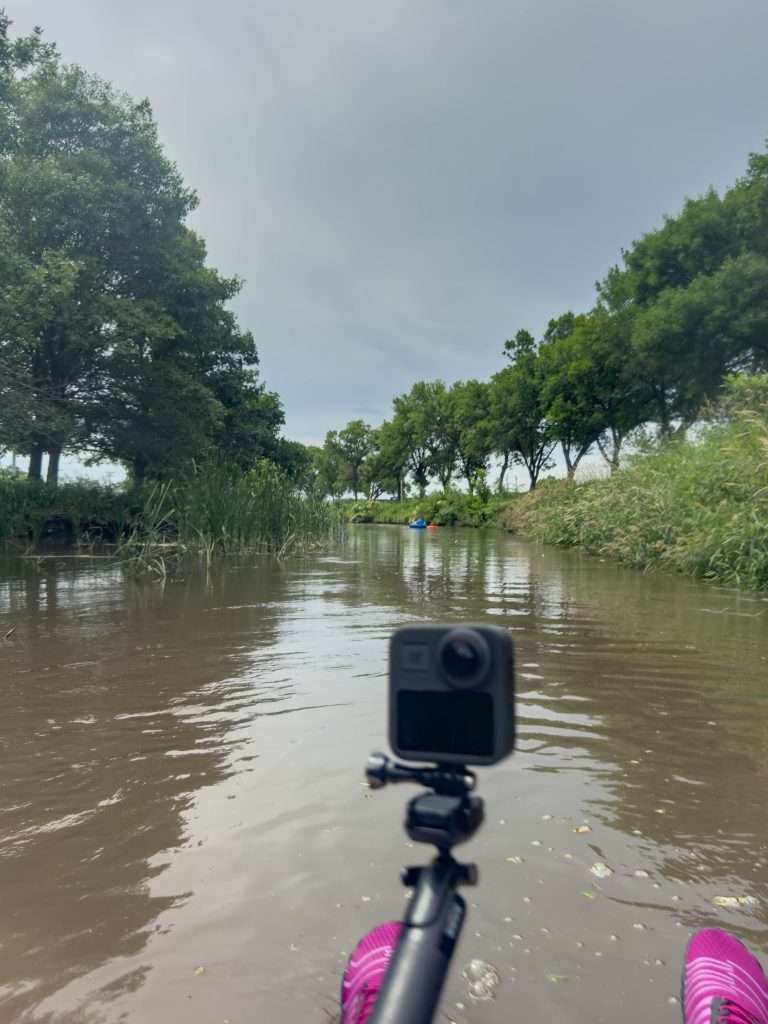 View from a river tube on the North Fork River in Norfolk, Nebraska, showing a mounted GoPro camera and pink water shoes with trees lining the waterway.