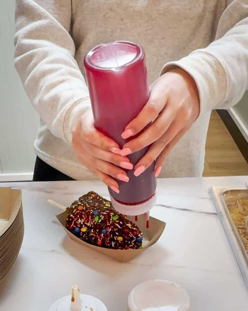 Employee drizzling fruit topping over a custom chocolate-covered frozen banana at Mythic Beach Banana Bar in Gulf Shores, Alabama