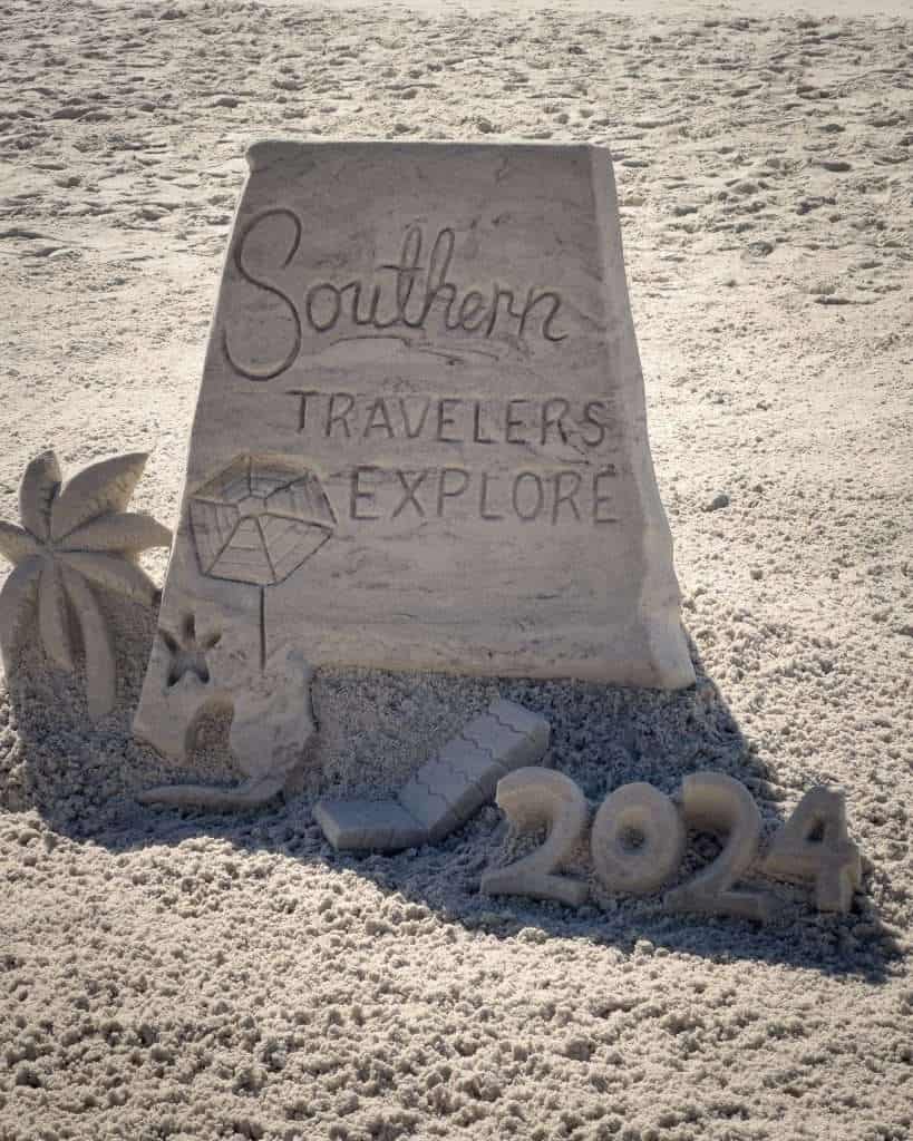 Sand sculpture on Gulf Shores beach with Southern Travelers Explore message, umbrella, starfish, and year 2024, celebrating beach travel and vacation fun.