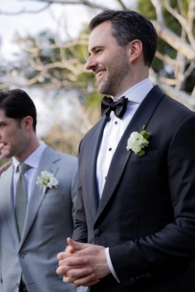 Groom in a classic black tuxedo with a white boutonniere, smiling during an outdoor wedding ceremony.