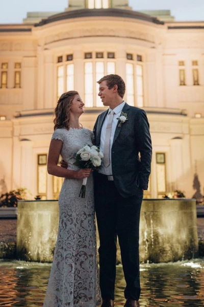Elegant wedding couple standing by fountain outside historic building at sunset.
