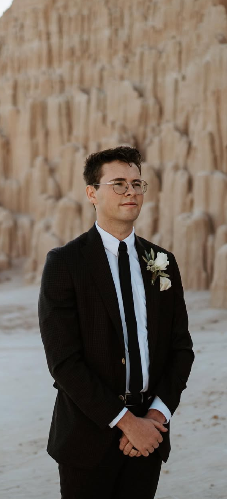 Groom in a black suit and glasses at a beach wedding with rocky cliffs in the background.