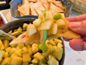 A hand holds a cinnamon sugar tortilla chip topped with diced green apples over a bowl of apple salsa.