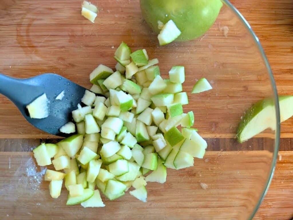 A glass bowl on a wooden surface containing a diced green apple and a black spatula.