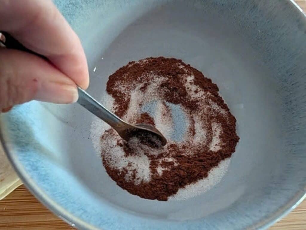 A spoon stirs a mixture of coffee grounds and sugar in a bowl.