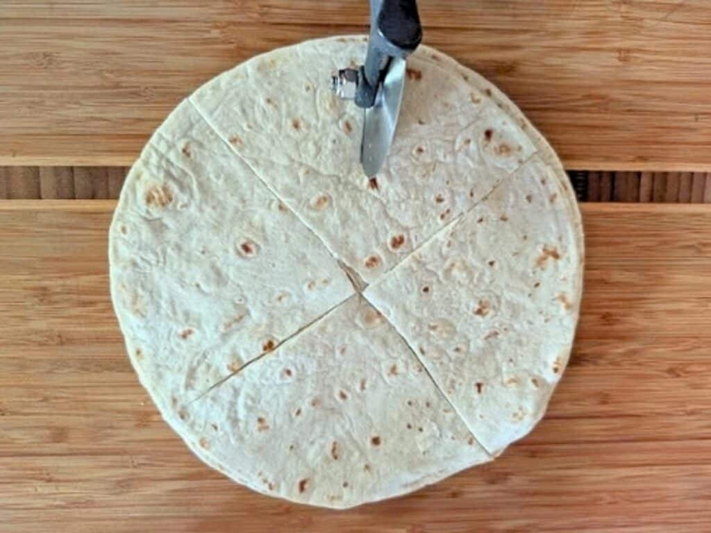 A circular tortilla being cut into four pieces with a pizza cutter on a wooden surface.
