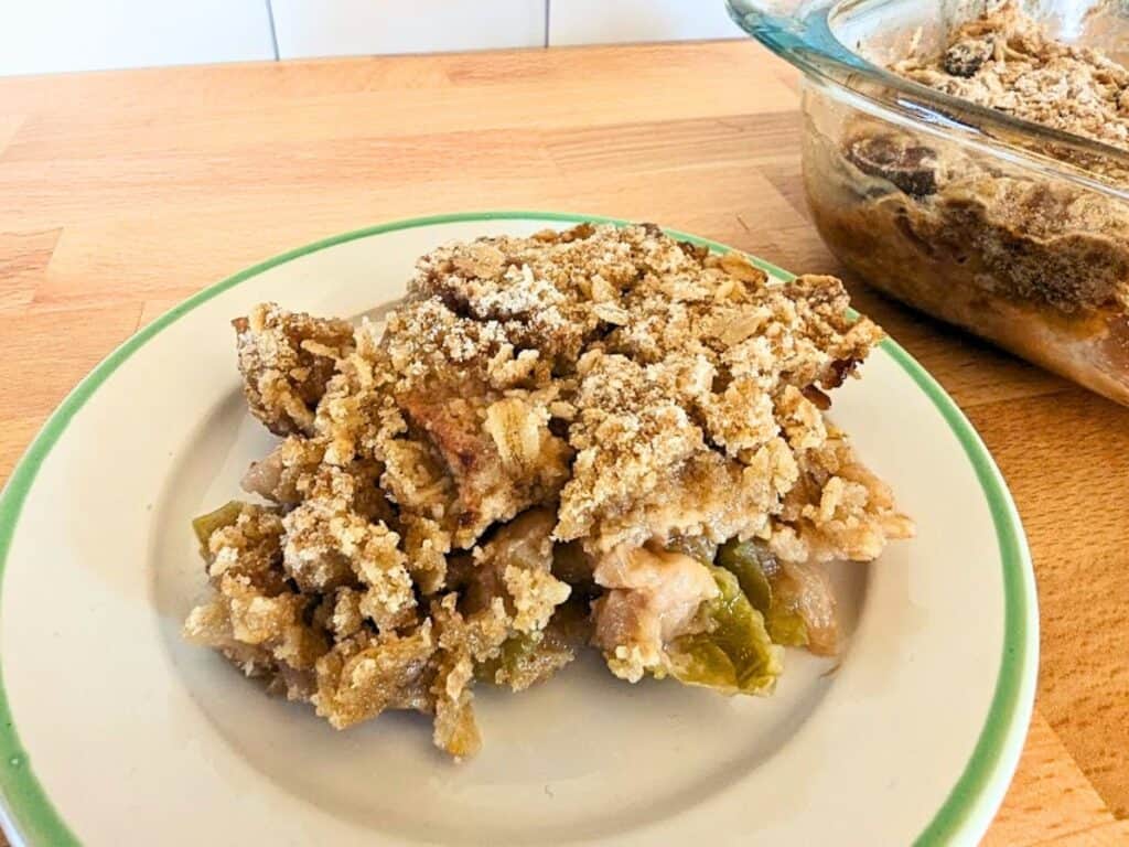 A serving of apple crumble with a crumbly topping is on a plate, with the rest of the dessert in a glass dish in the background on a wooden surface.