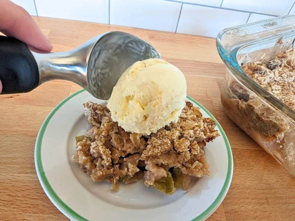 A scoop of vanilla ice cream is being placed on top of a serving of fruit crumble on a white plate, next to a baking dish.