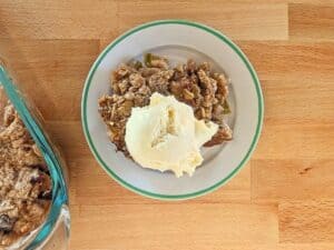 A plate with apple crumble topped with a scoop of vanilla ice cream sits on a wooden surface next to a glass baking dish.