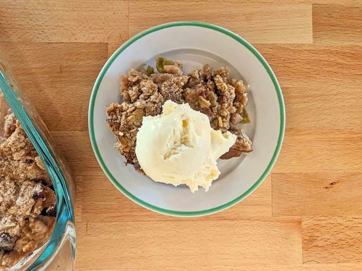 A plate with apple crumble topped with a scoop of vanilla ice cream sits on a wooden surface next to a glass baking dish.