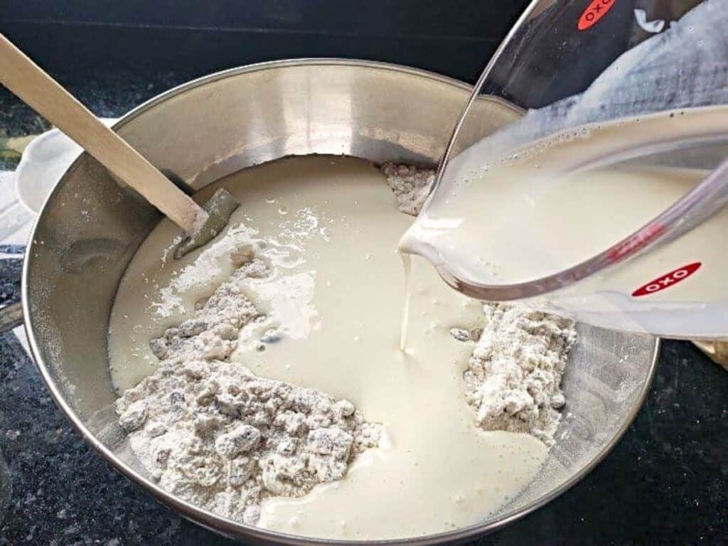 Milk is being poured from a measuring cup into a metal bowl containing flour, with a spatula resting on the side.