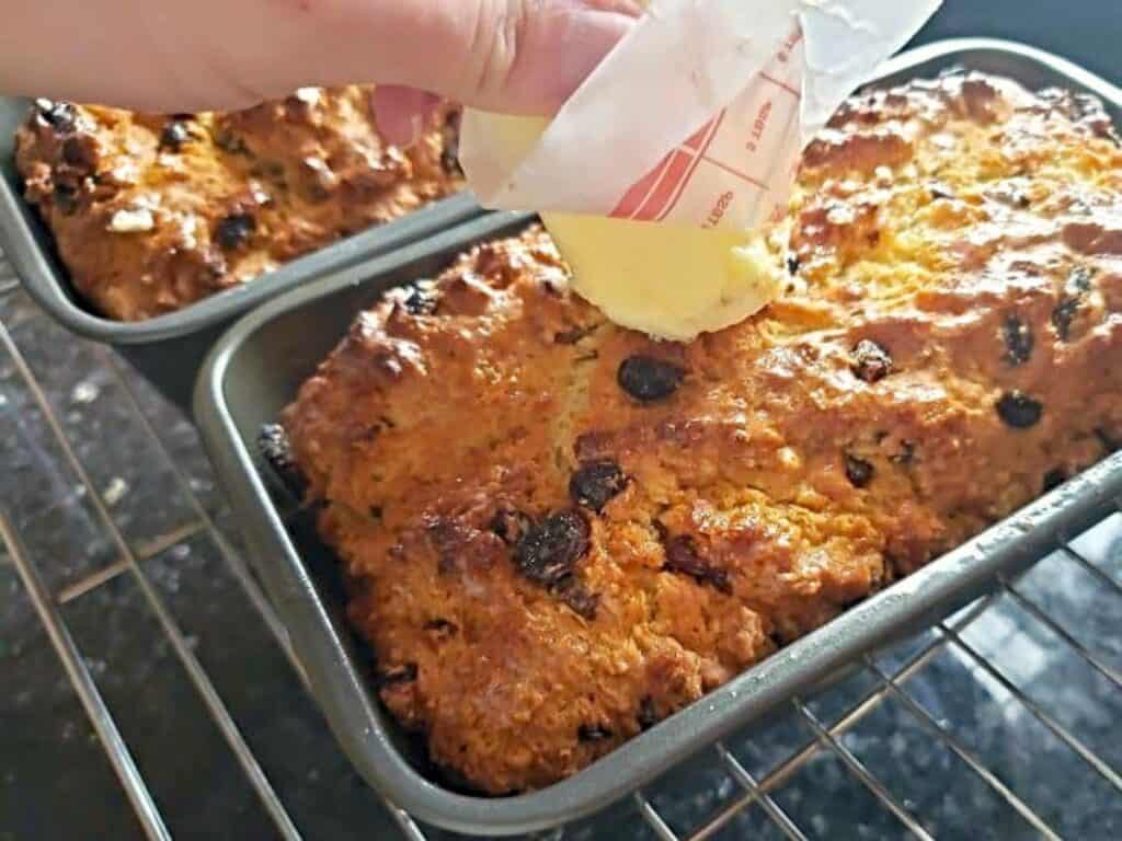A hand rubs a stick of butter over the top of freshly baked loaves with raisins in metal pans on an oven rack.