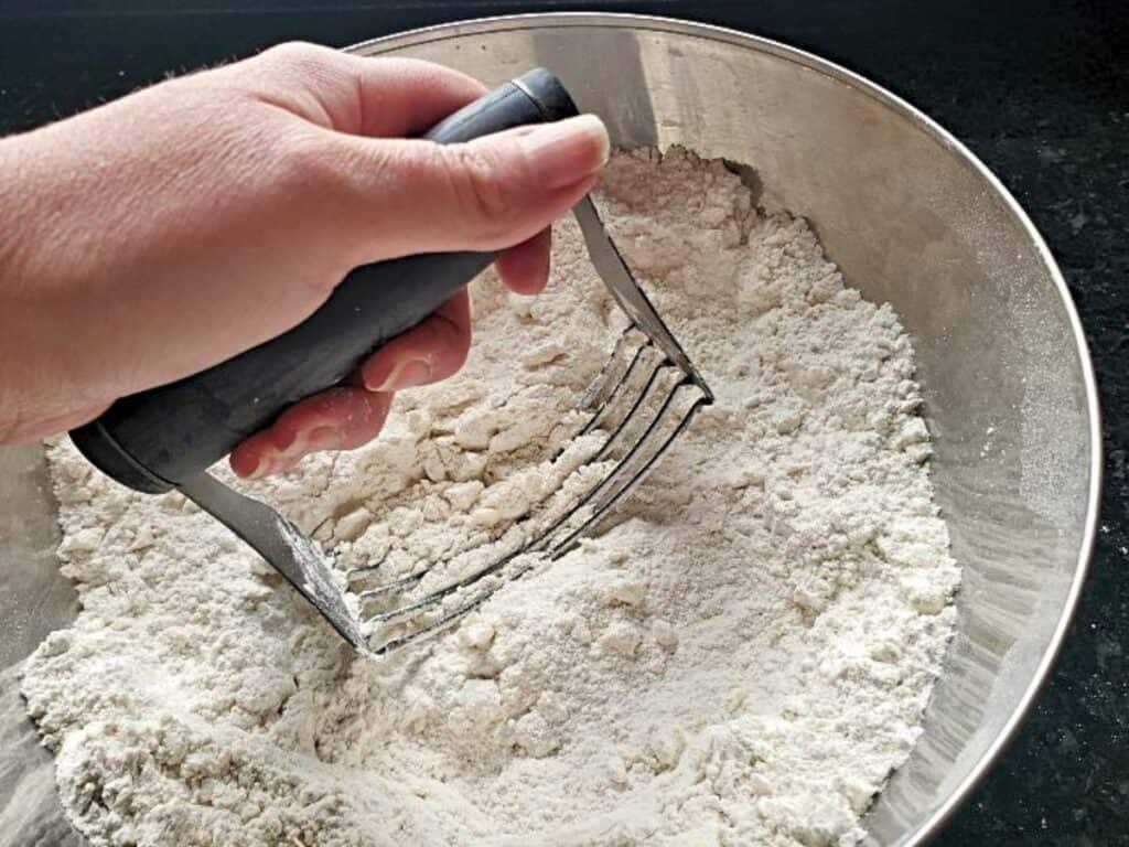 A hand uses a pastry blender to mix flour in a metal bowl.