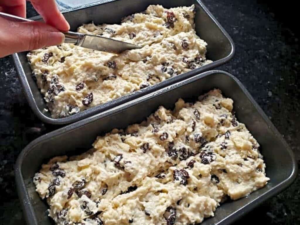 Two metal loaf pans filled with unbaked raisin bread batter; a hand uses a knife to score the top of one loaf.