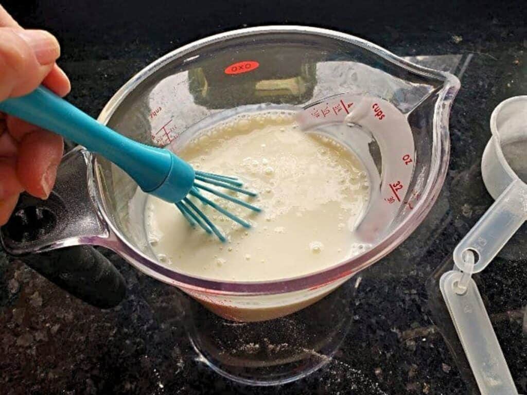A hand uses a blue whisk to mix a white liquid, likely milk, in a clear measuring cup on a dark countertop.