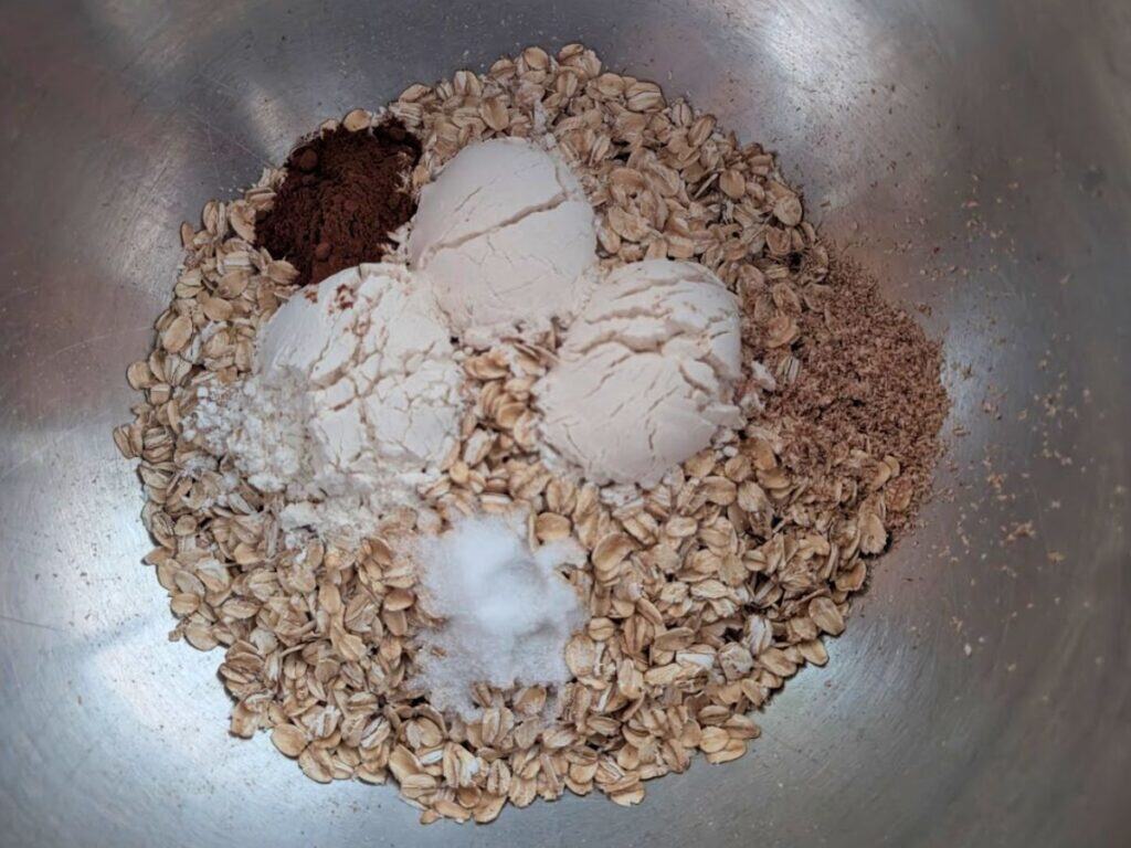 A metal bowl containing oats, flour, cocoa powder, ground flaxseed, salt, and baking soda—perfect for making delicious lacy cookies.