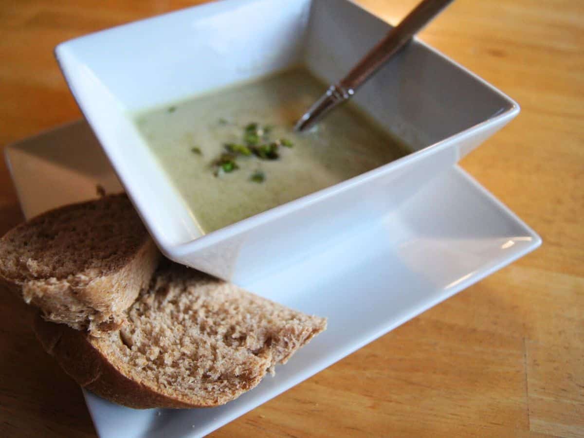 Image shows a square bowl of creamy green soup garnished with herbs, placed on a white plate with two slices of brown bread, and a spoon in the soup, on a wooden table.
