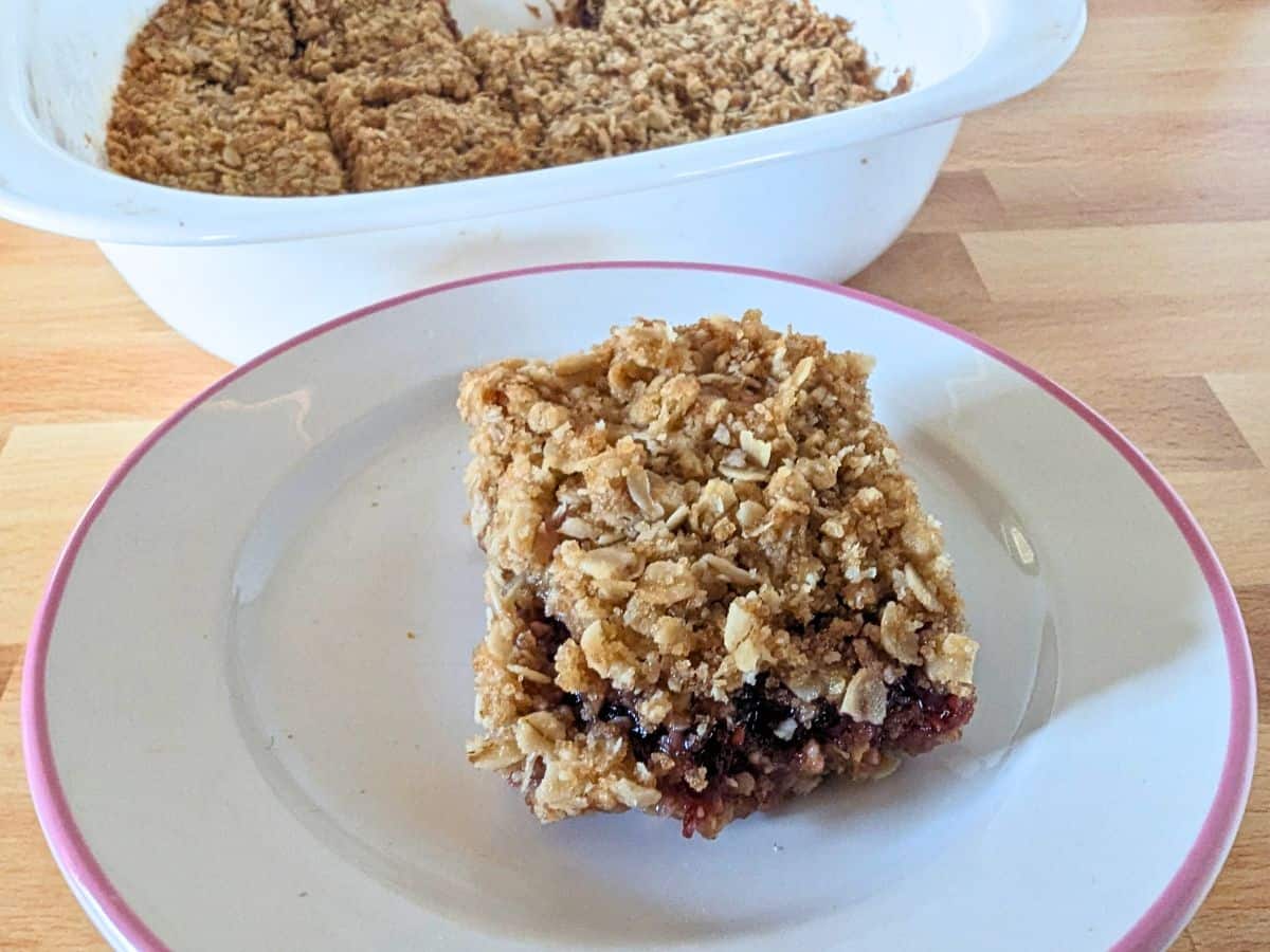 A square of a berry jam bar sits on a white plate with a pink rim. A white baking dish with the remaining crumble is in the background on a wooden surface.