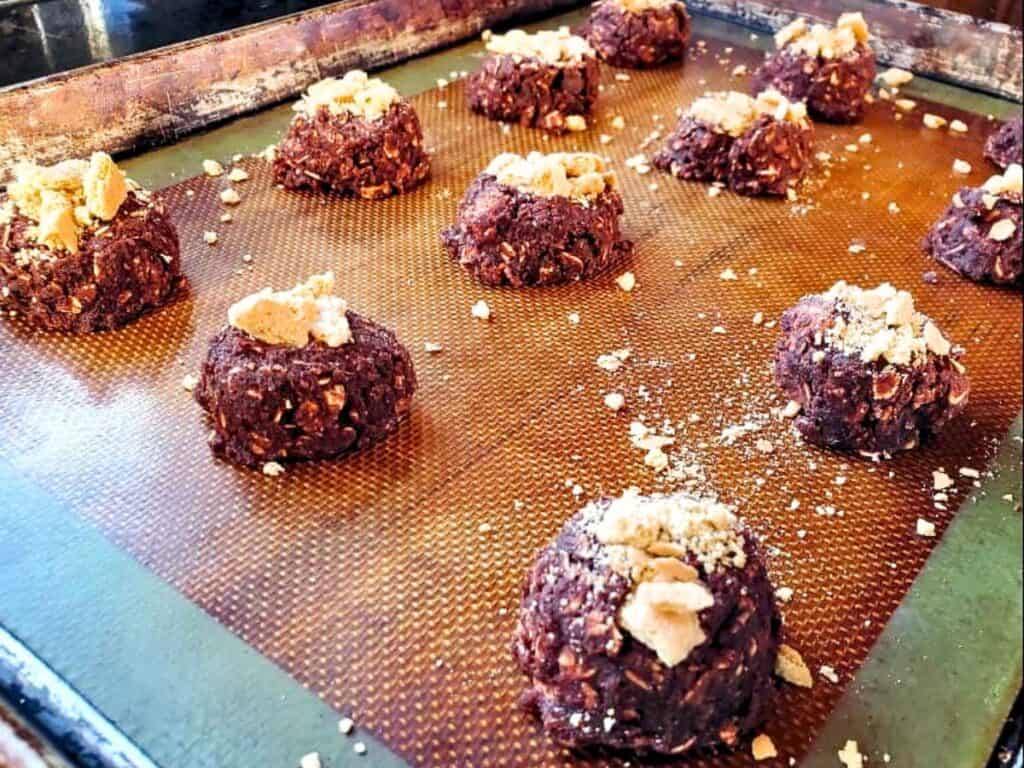 Unbaked chocolate cookie dough balls with oat flakes and a crushed topping sit on a silicone baking mat on a metal tray, ready to bake into chewy s’mores cookies.