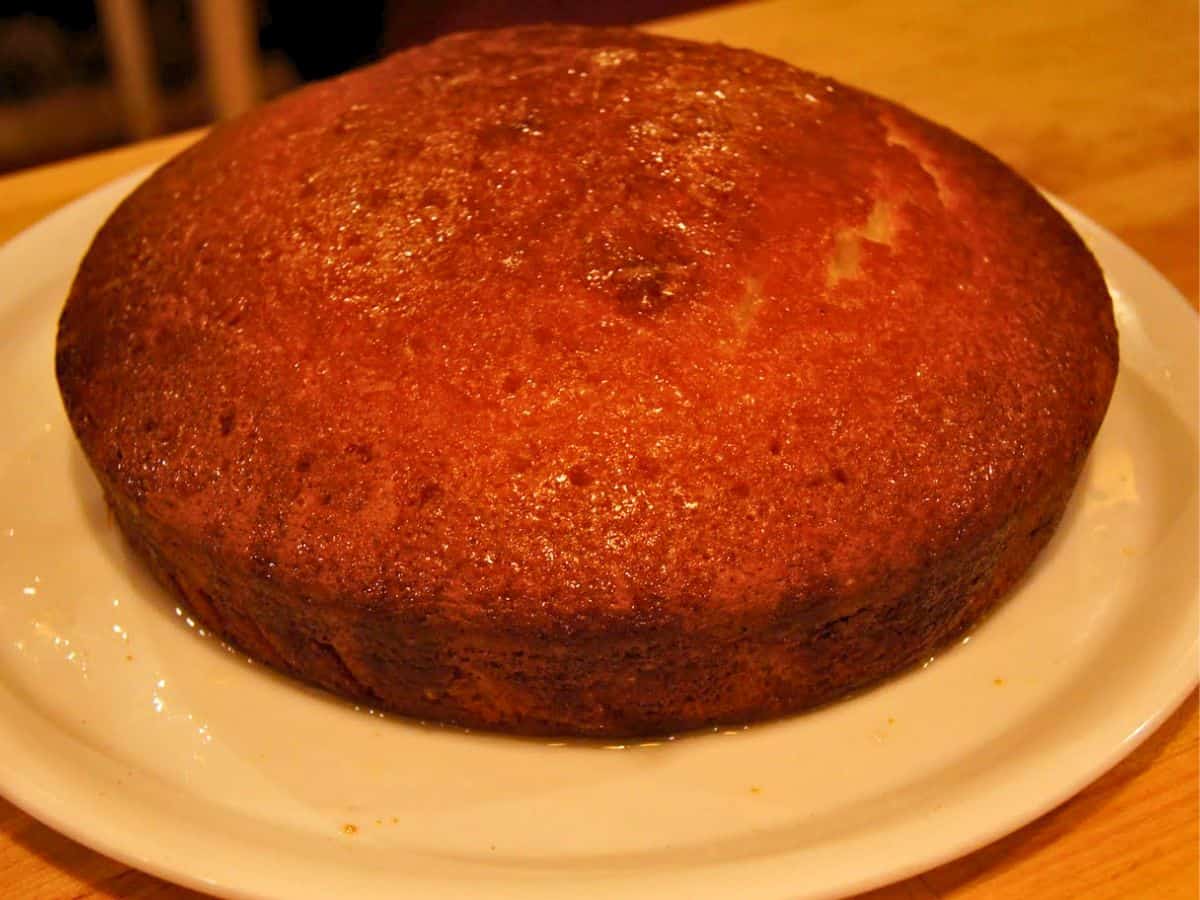 A round, golden-brown sponge cake sits on a white plate atop a wooden surface.