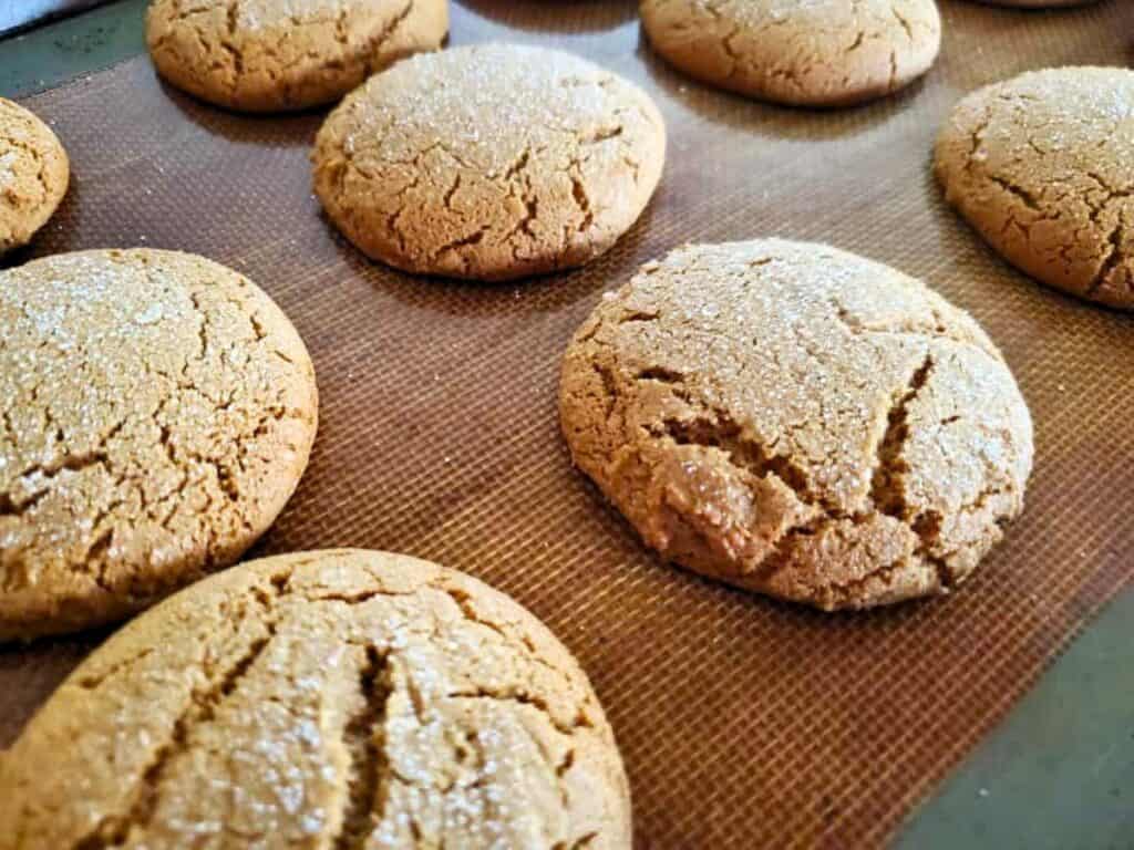 Close-up of several round, golden-brown chewy molasses cookies with cracked tops and a sprinkle of sugar, resting on a silicone baking mat.