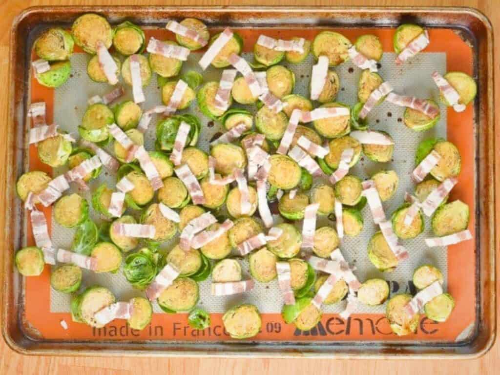 Halved Brussels sprouts and sliced bacon pieces are spread on a baking sheet lined with a silicone mat, perfect for making delicious bacon roasted Brussels sprouts.