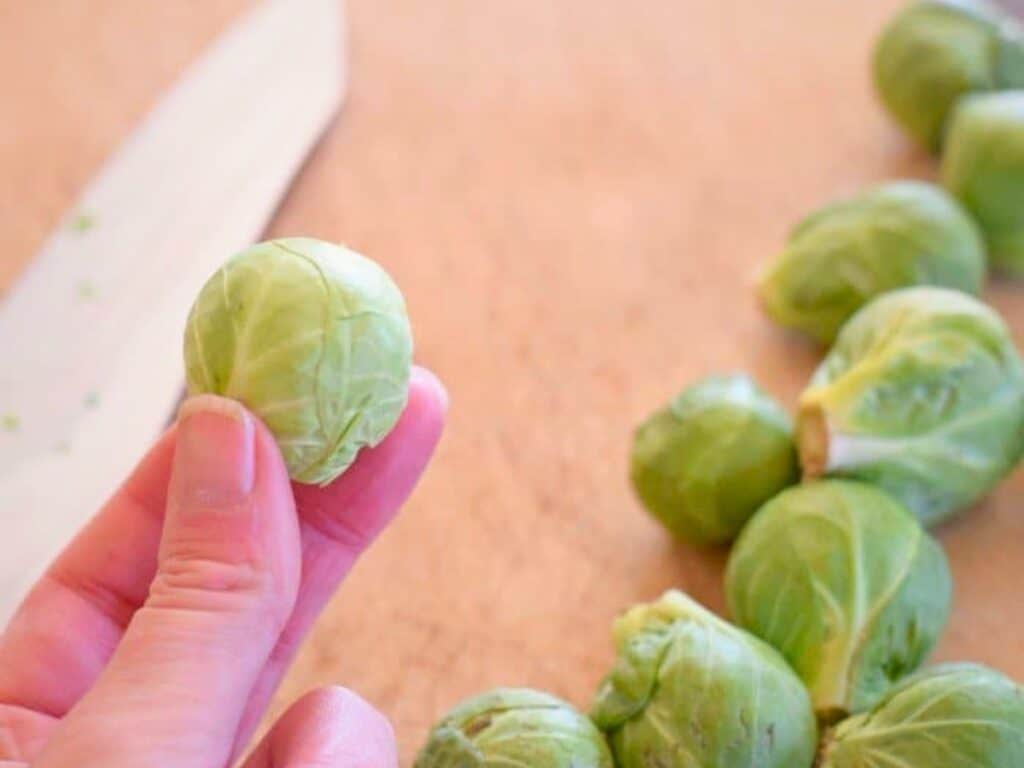 A hand holds a Brussels sprout over a cutting board, preparing for bacon roasted Brussels sprouts, with more Brussels sprouts and a knife in the background.