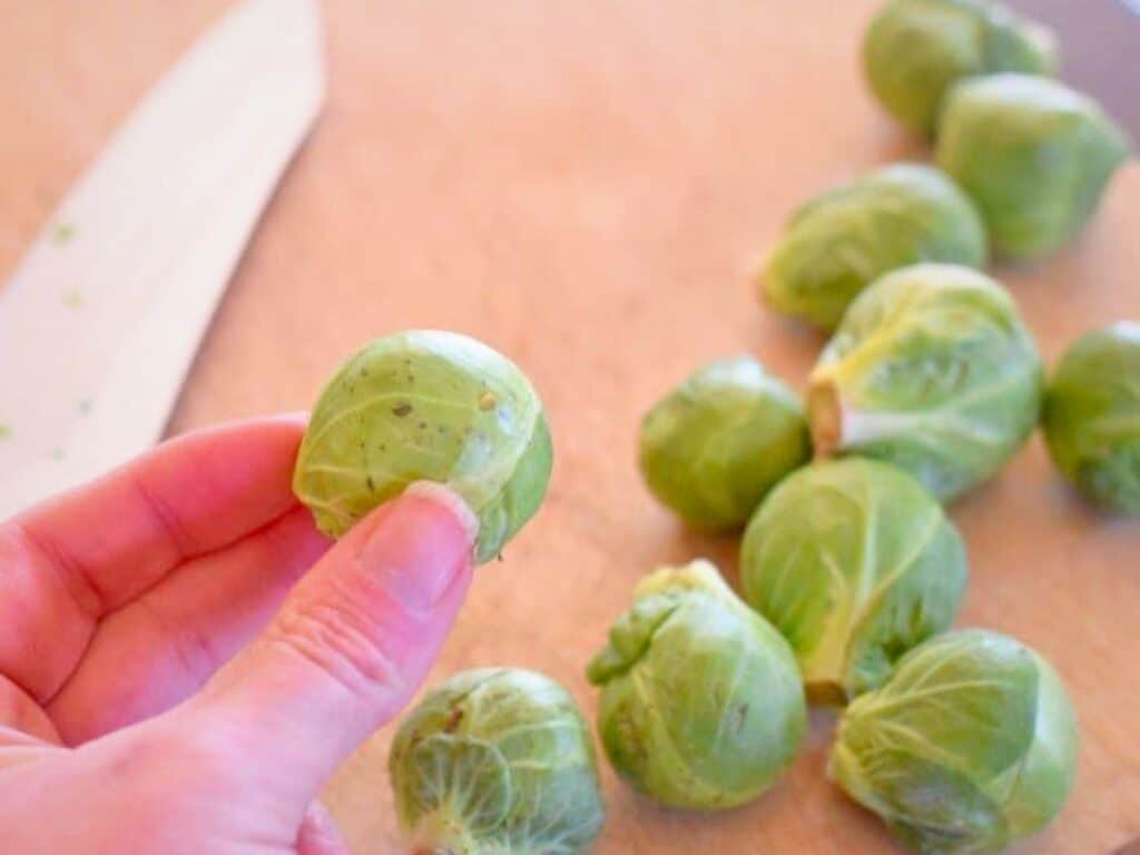 A hand holds a Brussels sprout over a cutting board with several other Brussels sprouts and a knife in the background, ready to be prepped for delicious bacon roasted brussels sprouts.
