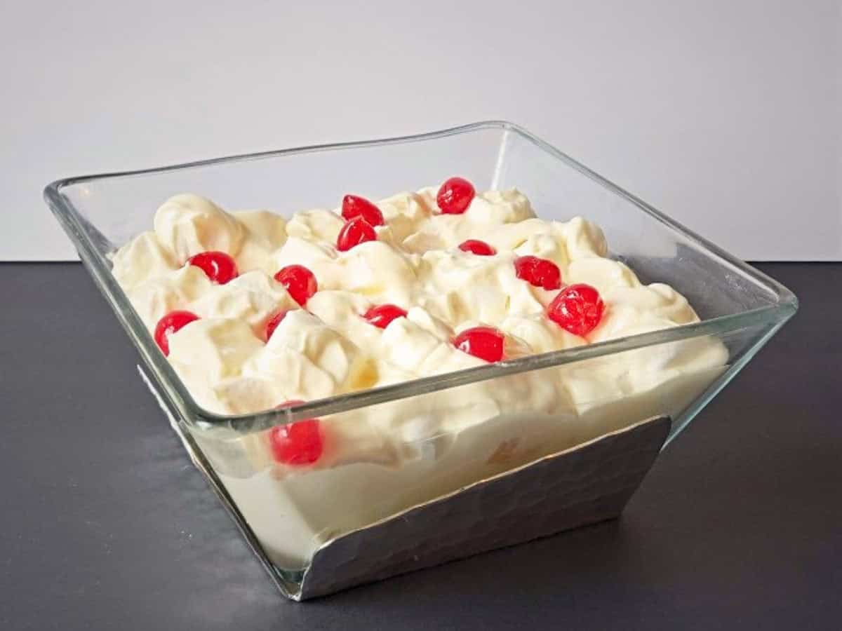 Glass bowl filled with creamy marshmallow salad, topped with several red maraschino cherries, set against a plain background.