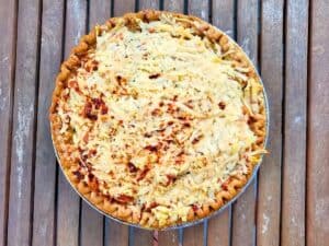 A baked savory pie topped with shredded cheese in a foil tin on a wooden surface.