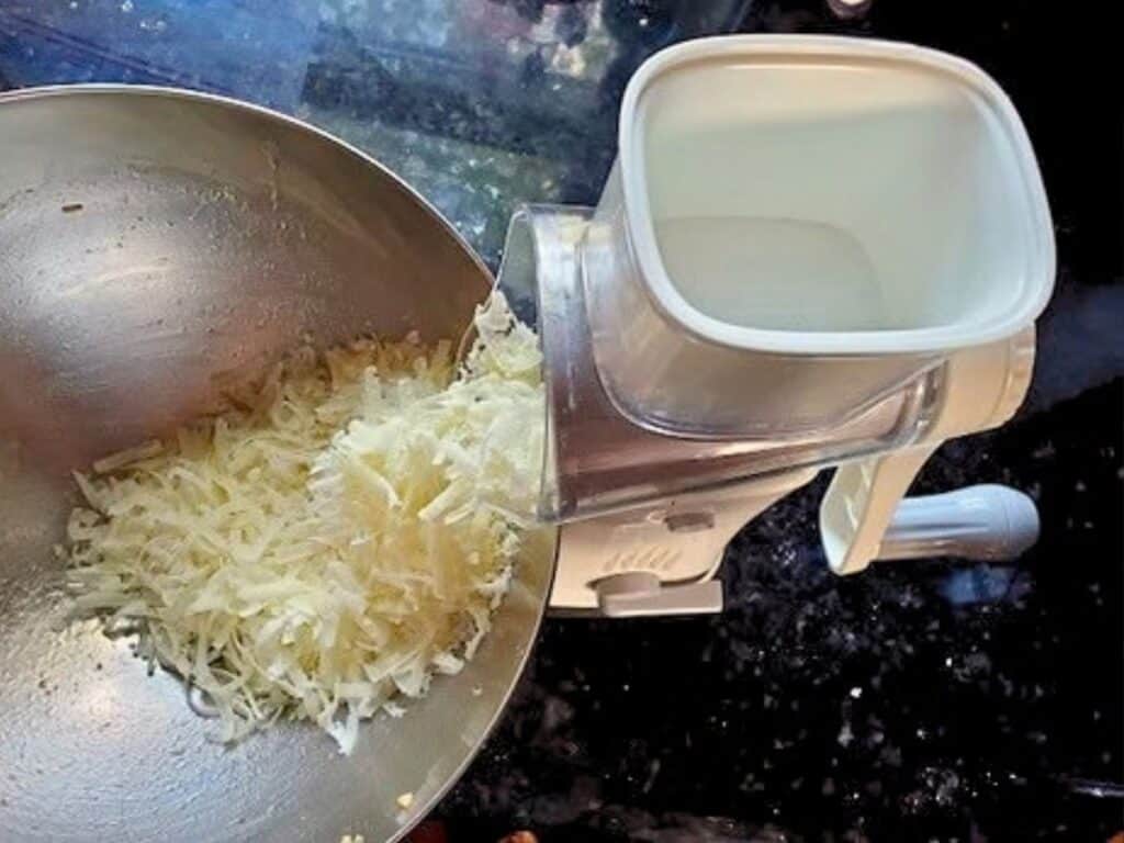 A hand-cranked rotary grater shredding cheese into a metal bowl.