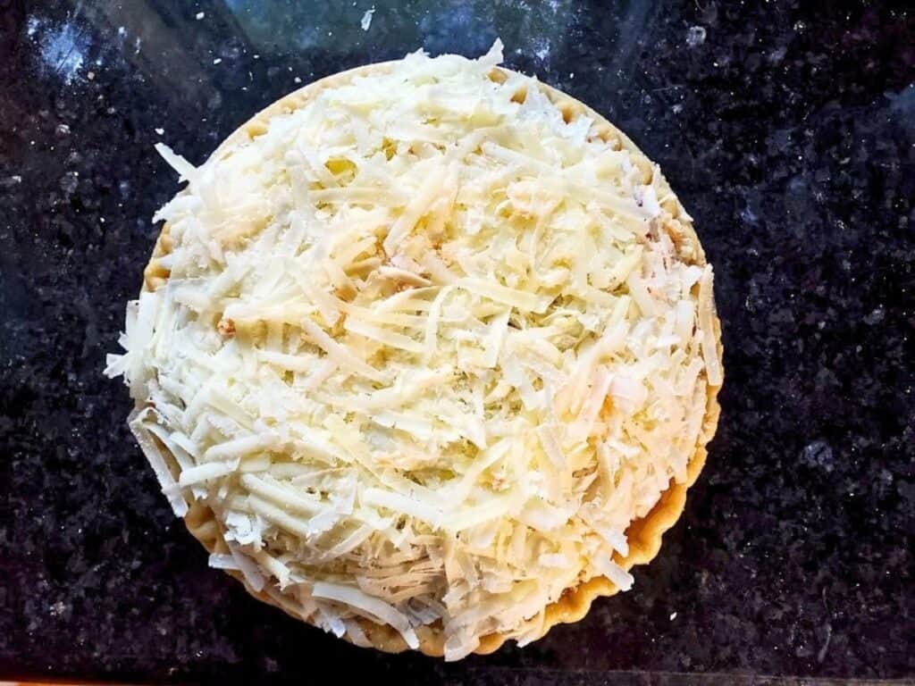An unbaked savory pie topped with a thick layer of shredded cheese.