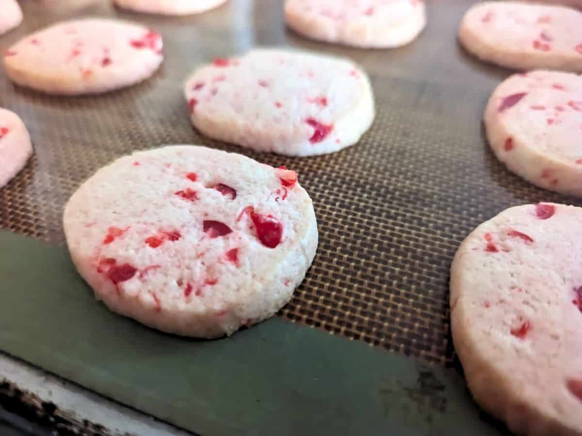Image shows round cookies with red berry pieces are lined up on a baking mat.