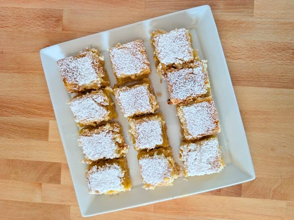 Twelve square pieces of baked lemonade cake topped with powdered sugar are arranged neatly on a white square plate, set on a wooden surface.
