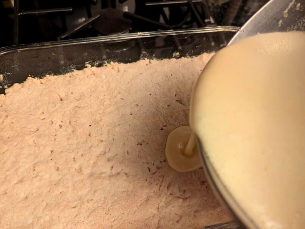 A bowl of liquid batter, inspired by baked lemonade, is being poured over a layer of crumbly dough in a glass baking dish on a stovetop.
