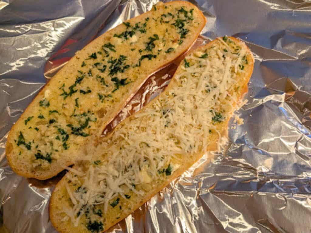 Two slices of bread on foil, one topped with garlic and herbs, the other with garlic, herbs, and shredded cheese, ready to be baked.