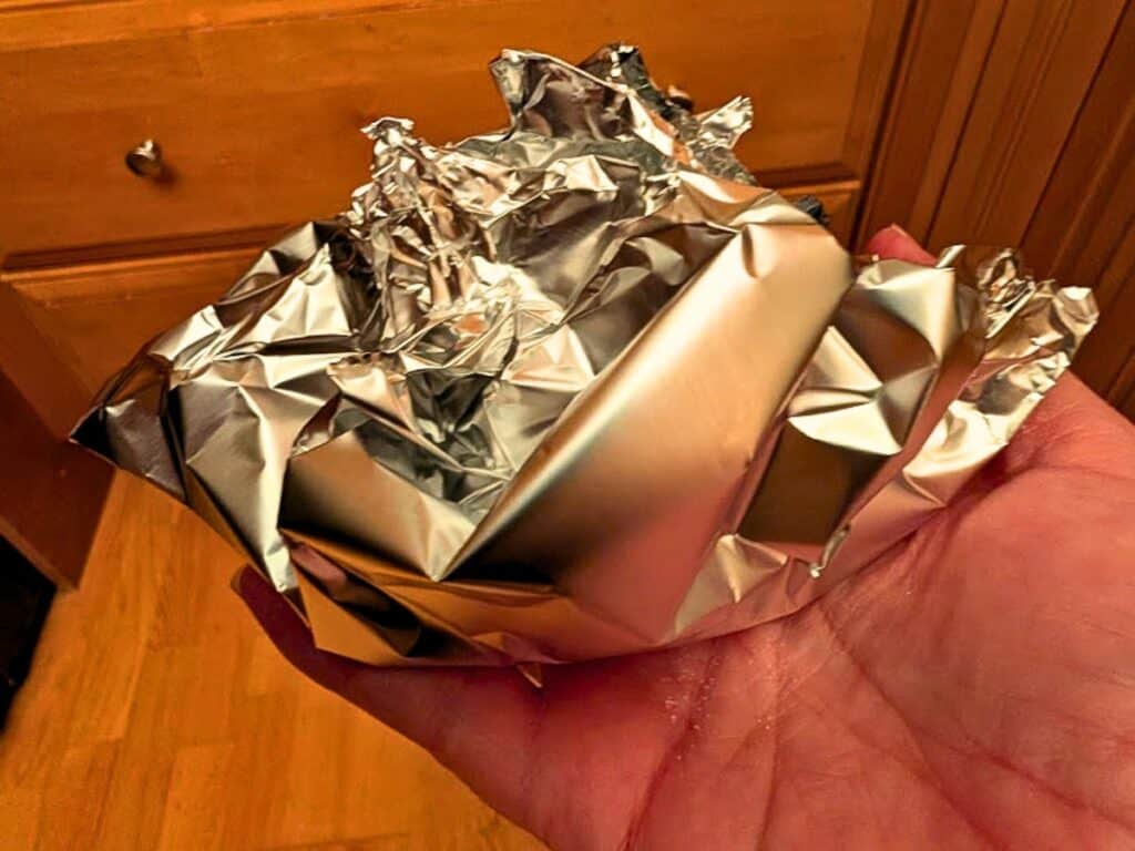 A hand is holding a crumpled sheet of aluminum foil indoors, with wooden drawers visible in the background.