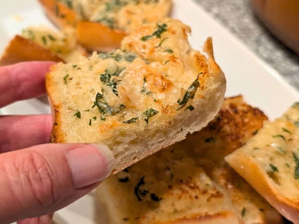 A hand holds a piece of garlic bread topped with melted cheese and chopped herbs, with more pieces on a white plate in the background.