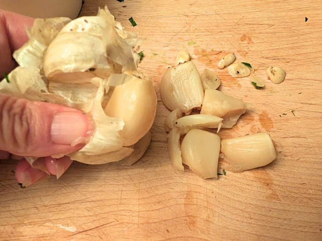 A hand peels garlic cloves on a wooden cutting board, with several peeled and unpeeled cloves visible.