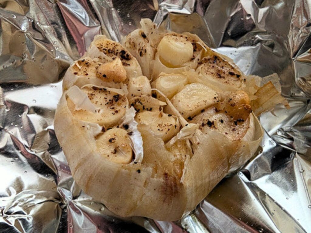 A roasted garlic bulb sits on crinkled aluminum foil, with cloves exposed and lightly browned, sprinkled with black pepper and seasoning.